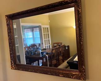 Ornate Gold Leaf Wall Mirror — This ornate gold leaf wall mirror features an intricately carved frame with detailed floral and scroll motifs. The large rectangular mirror is perfect for adding elegance and light to any dining room or living space. Measuring approximately 36 inches in height and 48 inches in width, it is a substantial decorative piece. The mirror and frame are in good condition with minor wear consistent with its vintage style. Its classic design makes it a timeless accent for traditional or vintage-inspired interiors.