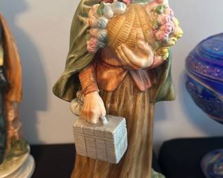Ardco Porcelain Lady Figurine — This charming Ardco porcelain figurine depicts an elderly lady carrying a basket of flowers and a picnic basket. Crafted with fine detail in warm earth tones, it features a green shawl, bonnet, and a flowing skirt that showcases skilled craftsmanship. The figurine stands approximately 9 inches tall, offering a delicate accent piece for any master bedroom or display cabinet. It is in good condition with minor signs of age and no visible chips or cracks. Made by Ardco in Dallas, this collectible porcelain piece reflects mid-20th century decorative style.