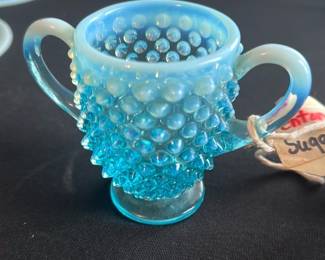 Blue Hobnail Glass Sugar Bowl — This charming blue hobnail glass sugar bowl features a distinctive textured pattern with raised knobs and a delicate opalescent gradient from light to deeper blue. The bowl includes two elegant handles and sits on a small round base, perfect for vintage or antique glassware collectors. Measuring approximately 3.5 inches in height and 4.5 inches in width, it fits nicely on a tea or coffee table. It is in excellent condition with no visible chips, cracks, or damage. This piece adds a touch of classic elegance to any master bedroom or dining set.