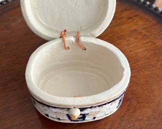 Ceramic Trinket Box — This charming Hughes Treasures ceramic trinket box features a sunflower design on the lid and a decorative floral band around its sides. The box has an oval shape and a hinged lid connected by leather cords. Measuring approximately 2.5 inches tall, 5 inches wide, and 4 inches deep, it provides a lovely small storage solution. The item is in good vintage condition with minor surface wear indicative of age. This collectible ceramic piece is perfect for storing jewelry or keepsakes, adding a rustic decorative touch to any room.