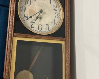 Oak Wall Regulator Clock — Oak wall regulator clock with carved surround and visible brass pendulum; a classic bedroom-up-room timepiece with ornate molded trim. The painted dial has large black numerals and decorative spade hands, set behind a glazed upper door and a glazed lower door showing the pendulum and winding key. Approximate overall size works for a hallway or parlor wall-mounted display and the movement appears to be a key-wind pendulum type (key included). Clock shows age: dial patina, some grime on face, light scuffs to wood and finish and the pendulum bob has surface wear; sold as-is for restoration or display.