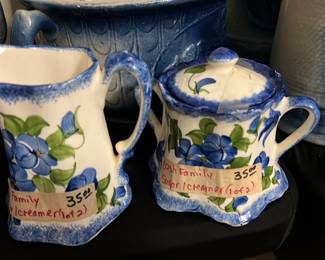 Clinahfeld Blue Floral Cream & Sugar — Pair of Clinahfeld (Clinahfeld Artware Pottery, Euvtn, Tenn.) blue floral ceramic creamer and covered sugar bowl from the Bonus Up Room. Both pieces have white glaze with hand-painted blue flowers and green leaves, blue sponged trim, and scalloped bases; sugar has a fitted lid. Overall small tableware size suitable for display or light use. Both show surface wear and residue on bases consistent with age and prior use but no obvious large chips or cracks visible in photos.