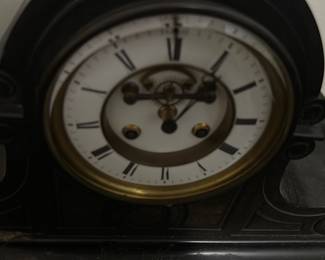 Antique Black marble Mantel Clock — This antique black mantel clock features a curved body with elegant scroll details, showcasing classic early 20th-century design. The clock face has Roman numerals and visible winding holes, framed by a brass bezel with an enamel dial. Measuring approximately 10 in high, 18 in wide, and 6 in deep, this clock is an authentic decorative timepiece ideal for a vintage mantel or shelf. It shows signs of surface wear and dust consistent with age but appears structurally sound. This piece is a fine example of traditional clockmaking, adding nostalgic charm to any bedroom or living room setting.