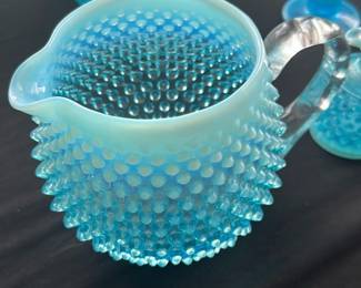 Blue Hobnail Milk Glass Pitcher — This charming blue hobnail milk glass pitcher features a textured bubble design with a clear twisted handle for easy pouring. The pitcher exhibits a beautiful opaline blue color with classic vintage appeal. Measuring approximately 5.5 inches tall and 6 inches wide including the handle, it makes an elegant accent for any tabletop or display. The piece is in good condition with no visible chips or cracks, showing minor wear consistent with vintage glassware. Its distinctive hobnail pattern and opaline glass make it a collectible item for vintage glass enthusiasts.
