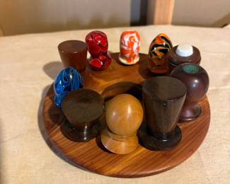Collection of handcrafted turned wood pieces including bottle stoppers, decorative knobs, miniature vessels, and colorful acrylic woodturning creations displayed on a matching wood stand.