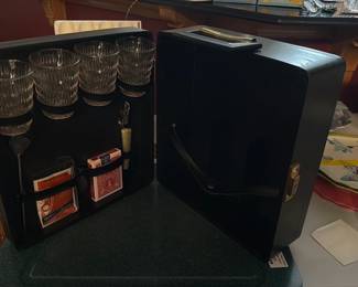Antique Portable Black Cocktail Bar Case — Compact black portable cocktail bar case with molded handle and interior fitted to hold four cut glass tumblers, two decks of playing cards, a spoon, and a small corkscrew/foil cutter. The exterior is a simple black hard case with brass-tone latch and plastic handle; interior has elastic straps and molded slots to secure glassware and accessories. Measures approximately 12 in tall, 10 in wide and 4 in deep (small and easy to carry for travel or picnics). In good used condition with light scuffs to exterior and clean interior; glasses appear intact with no visible chips.
