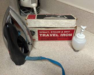 Vintage GE Travel Iron F49 — Vintage GE spray, steam & dry travel iron (model F49) in black with chrome soleplate, shown with original box in laundry room. Compact sprung-handle travel design with blue cord and small refillable spray bottle; intended for 120 or 250 volts AC according to box. Measures approximately 6.5 inches tall, 3.5 inches wide and 8 inches long — compact for travel use. Shows surface wear and scuffs on soleplate and cord sleeve; functional condition untested so sold as-is for parts or restoration.