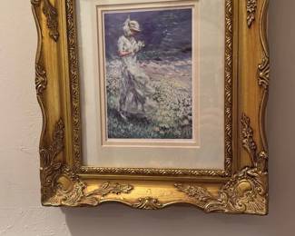 Ornate Gold Framed Prints (Pair) — Pair of floral and seaside art prints in matching ornate gold frames with decorative molded corners and a double mat surround. Each piece features a pastel still-life vase composition and a woman walking on a flowered shoreline, both with soft impressionistic brushwork. Frames measure approximately 20 in tall by 16 in wide and are relatively lightweight wall-hangings suitable for a bedroom or hallway. Both are in good condition with minor surface scuffs to the frames and intact glass and mats.