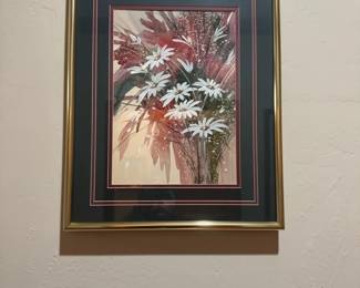 Floral Print Wall Art in Gold Frame — This framed floral print features an elegant arrangement of white daisies set against a soft, muted background with warm red and pink tones. The piece is accented with a classic gold metal frame and dual colored black and maroon matting, adding depth and contrast to the artwork. Measuring approximately 19 inches in width and 26 inches in height, it is an ideal size for accenting a variety of interior spaces. The print is protected behind glass and the frame is in good condition with minor signs of age but no significant damage. Perfect for adding a touch of floral charm to any room, this framed print complements traditional and contemporary decor styles.