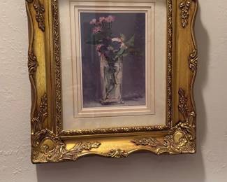 Ornate Gold Framed Prints (Pair) — Pair of floral and seaside art prints in matching ornate gold frames with decorative molded corners and a double mat surround. Each piece features a pastel still-life vase composition and a woman walking on a flowered shoreline, both with soft impressionistic brushwork. Frames measure approximately 20 in tall by 16 in wide and are relatively lightweight wall-hangings suitable for a bedroom or hallway. Both are in good condition with minor surface scuffs to the frames and intact glass and mats.