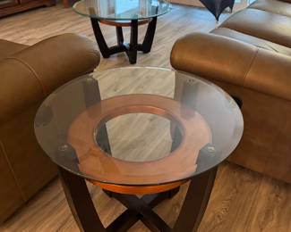 Add a sophisticated contemporary touch to your living room with this stunning three-piece occasional table set. The collection features a round tempered glass coffee table and two matching end tables, each showcasing a striking architectural base with black wood supports and a warm circular wood accent beneath the glass.