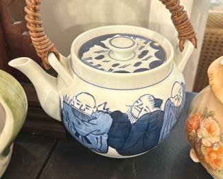 Blue & White Ceramic Tea Pot — Hand-painted blue-and-white ceramic teapot with woven wicker handle and matching lid. The body features stylized figures and botanical motifs in deep cobalt underglaze; the lid echoes the pattern. Handle shows intact wrapping and the pot appears structurally sound with only light surface wear consistent with display use. A decorative, functional piece ideal for serving or display; measures roughly as listed below.