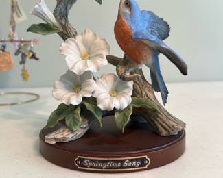Springtime Song Porcelain Birds — Ornate porcelain figurine titled "Springtime Song" featuring two blue-and-orange songbirds perched on a flowering branch with detailed leaves and blossoms on a wood-tone oval base. The piece displays fine molded detail and soft hand-painted colors typical of collectible decorative porcelain; the small brass-look nameplate reads "Springtime Song." Overall condition appears good with no visible chips or cracks in photos. A compact decorative accent for a mantel, shelf, or curio cabinet measuring roughly a foot tall.