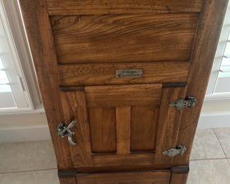 Gibson's Cambria Oak Icebox — This Gibson's Cambria oak icebox is a charming antique-style wooden refrigeration cabinet with original metal nameplate and decorative hardware. Constructed of solid oak with a painted white insulated interior and removable wire shelf, it features a top-opening ice compartment and a lower storage door. Overall footprint is modest and practical for a sun room or kitchen as an accent piece; measurements provided are estimated. Condition is good structurally with surface wear, scuffs to interior paint, and signs of age on metal fittings consistent with regular use.