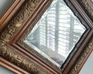 Ornate Wood & Gilt Wall Mirror — Decorative ornate wall mirror with carved wood frame and gilt-accented inner molding, photographed in the living room. The frame features layered moldings with floral and scroll relief details and a beveled inner mirror; finish is warm wood tone with gold highlights. Approximate overall footprint is moderate-sized and ideal for entryway or living room accent; lightweight enough for standard wall mounting. Mirror shows slight age-related spotting on the glass and normal surface wear to the frame consistent with prior use.