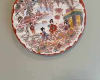 Decorative Chinese Famille Rose Plate — Decorative famille rose style porcelain plate with scalloped rim and hand-painted scene of figures in a garden. The plate features pink, red, green and yellow enamels with gilt highlights and a red scalloped border; photographed hanging in master bedroom. Approximate measurements and weight are estimates; plate is in good condition with no visible cracks or chips from photos but shows minor surface wear consistent with display use. A charming decorative wall plate suitable for display or collection.