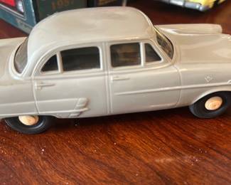 Gray Classic Toy Car — Gray diecast toy sedan model displayed in office room; solid metal body with chrome-plated bumpers and plastic wheels. Clean lines and hood ornament detail suggest a 1950s-style American sedan replica; paint shows light surface wear and a few scuffs. Approximately palm-size and lightweight, suitable for display or a collector of classic toy cars. Condition is good with expected play/age wear and intact wheels and bumpers.