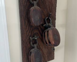 Wooden Pulley Hook Wall Display — Decorative wall-mounted display of three reclaimed wooden pulley blocks mounted on a distressed plank, ideal for nautical or rustic sun room decor. The pulleys are solid wood with metal swivel hardware and decorative brass medallions; the plank shows deep grain and weathered patina. Overall mount is compact and light enough for easy hanging (see dimensions) and works well as an accent in a sun room or entry. Good vintage condition with expected wear, surface scratches, and patina consistent with age and prior use; hardware appears intact though aged.
