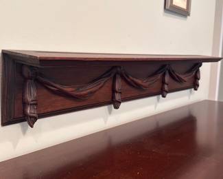 Carved Wood Wall Shelf — Solid carved wood wall shelf with swag and tassel motif across the front; the shelf has a classic traditional look and would work as a small display ledge or mantel accent. Construction appears to be stained hardwood with molded top edge and three carved swag/tassel brackets; there is light surface wear and small edge chips consistent with normal use. Mounting keyholes are visible on the underside for wall installation; depth is shallow so it suits small picture frames or decorative objects. Attractive carved detail gives it decorative appeal while remaining practical for entry, hallway, or bedroom use.