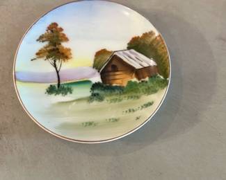 Hand Painted Made in Occupied Japan — Small hand-painted porcelain wall plate marked "hand painted" and "MADE IN OCCUPIED JAPAN" on the back. The front shows a pastoral scene with a small cabin, tree and soft landscape in muted greens, browns and lilac tones with a thin gilt rim. Piece is a collectible souvenir-era porcelain likely from the late 1940s; photos show light surface wear and the glaze intact with no obvious chips or cracks. Includes original hanging wire on the back; diameter and weight are estimates below.