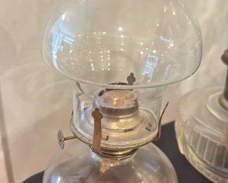 Glass Oil Kerosene Lamp — Small glass oil/kerosene lamp with clear glass chimney and round reservoir base showing residual amber residue inside. Metal burner assembly with wick-raising knob and fabric wick intact; burner shows surface rust and patina consistent with age. A compact decorative or functional piece for collectors of lighting; sold as-is with cosmetic wear and residue inside the base.