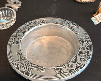 Pewter Wine Bottle Coaster — Heavy decorative pewter round dish with a shallow bowl center and a wide floral-embossed rim. The piece appears solidly made with a matte aged patina and light surface wear consistent with use and age; no obvious breaks or repairs visible in the photos. Good for holding keys, jewelry, or as a decorative accent on a dresser or entry table. Measures compactly and feels substantial in hand.
