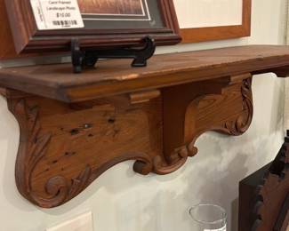Carved Wooden Wall Shelf — Carved wooden wall shelf with scrollwork and central bracket support, shown mounted and photographed from multiple angles. The piece features hand-carved foliate and scroll details on solid wood with a warm medium-brown finish and visible nail holes and surface wear consistent with age. Useful as a small display ledge for framed art or collectibles; sturdy-looking but sold as-is with cosmetic dings and finish wear shown in photos. Overall depth is shallow—good for lightweight decorative items.