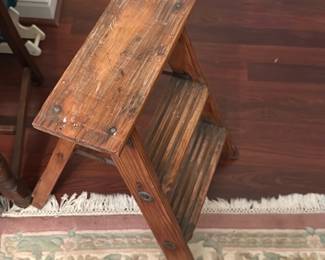 Wooden Two-Step Folding Stool — Solid wooden two-step folding stool with original patina and visible wear consistent with regular use. Constructed from slatted boards with metal hinge hardware, it folds flat for storage and would work well as a rustic step or plant stand. Shows surface scratches, old paint flecks, and scuffs but remains structurally sound. Compact footprint makes it useful in a kitchen, workshop, or bonus room.