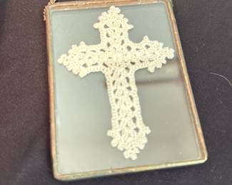 Framed Crochet Cross Wall Hanging — Small rectangular mirror with a pressed-lace cross mounted behind glass in a thin metal bezel and short hanging chain. The piece appears handmade or vintage folk-style with light surface wear to the metal frame and minor spotting on the mirror visible in photos. Ideal as a devotional wall accent or small decorative mirror; measures compactly and is lightweight for easy hanging.