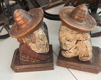 Carved Wood Mexican Hat Bookends — A pair of carved wooden bookends depicting robed figures wearing wide-brimmed Mexican-style hats. Each sits on a rectangular base with painted and carved surface detail showing intentional distressing and age-related wear; paint loss and surface chips are visible, especially on the robes and bases. Solid, decorative shelf pieces suitable for rustic or folk-art displays; measurements are approximate and condition is as photographed with noticeable paint loss and scuffs.