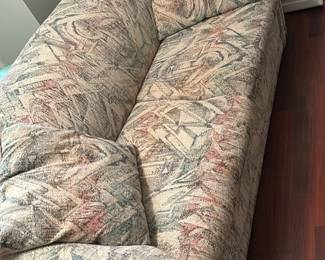 Patterned Upholstered Sofa — Three-seat patterned upholstered sofa with loose back and seat cushions and two matching throw pillows. The fabric shows a multicolor abstract/geometric print in muted pastels and the frame has low rolled arms; construction appears typical of late-20th-century casual living-room sofas. Cushions show visible wear and some flattening consistent with regular use but no obvious tears or stains in photos. Measures roughly like a standard sofa; good candidate for reupholstery or comfortable family-room seating.