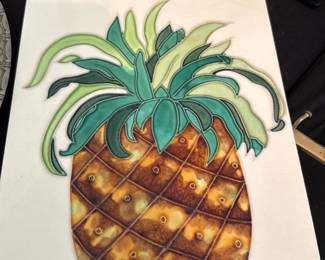 Hanging Tile Pineapple Wall Art — Decorative hanging tile featuring a raised, glazed pineapple motif in warm ambers and green leaves on a white ground. The tile is mounted on a fiberboard backing with cork edge and appears designed for wall display; photos show light surface crazing and typical age wear but no obvious breaks. A cheerful kitchen or entry accent; lightweight and easy to hang as shown.