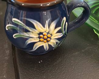 Hand-Painted Blue Creamer Pitcher — Small hand-painted ceramic creamer pitcher in a deep blue glaze with a floral motif and warm brown interior. The round form has a single handle and a spout for pouring; brushwork shows a stylized white-and-yellow flower with green leaves. Good condition with normal light surface wear visible in photos and no obvious chips or cracks. Compact size works well for tabletop use or decorative display.