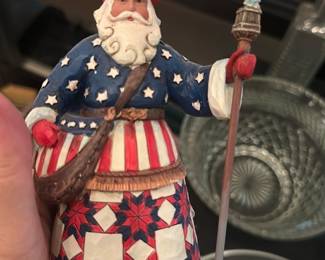 Jim Shore Patriotic Santa Figurine — Hand-painted Jim Shore Santa in patriotic red, white and blue attire, holding a staff topped with a tiny Statue of Liberty. The folk-art style features Jim Shore's characteristic quilted/folk patterns on the skirt and hand-painted floral border at the base. Useful as seasonal decor or a collectible for Jim Shore collectors; shows light surface wear consistent with display but no major chips or cracks visible in photos. Measures approximately 9 inches tall (see dimensions) and includes original resin painted finish.