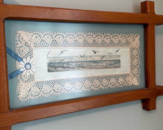 Framed Cross Stitch Wall Art — Decorative framed wall piece featuring a rectangular lace doily mounted over a pale blue mat with a narrow panoramic print of birds in flight at the center. The oak-toned wooden frame has a mid-century feel with simple corner extensions and the glass front appears intact; mat and lace are clean with minor age toning visible on close inspection. Lightweight, decorative wall art suited to bedroom or living areas—ready to hang as shown. Overall condition is good with normal surface wear to the wooden frame.