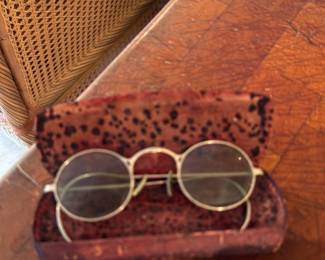 NYC Antique Eyeglasses — Small round wire-rim eyeglasses shown tucked into a fitted hard case with mottled red interior. Metal frames appear gold-tone with thin temples and round lenses suggesting an early 20th–mid century style or costume jewelry eyewear; lenses show light age-related haze and the case has wear to the covering. Useful as a wearable vintage accessory with minor cosmetic wear consistent with age.