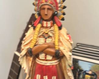 Wooden Native American Figure — Carved wooden figure of a Native American wearing a feathered headdress and traditional dress with painted detail in red, yellow and cream tones. The piece appears hand-carved with layered clothing and braids, standing in a relaxed pose with arms crossed; surface shows minor scuffs and light wear consistent with age and display. Good decorative condition with intact paint and no obvious cracks visible in the photos. Measures roughly tabletop size and works well as a mantel or shelf accent.