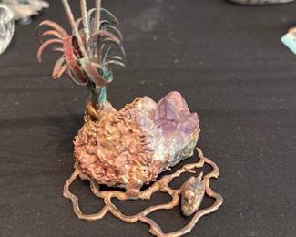 Amethyst & Metal Art Piece — Small decorative amethyst cluster mounted in a sculptural metal base with organic vine and plant motifs. The piece combines a natural purple crystal point attached to a textured rock base with a patinated metal frame and tiny bird detail; it reads as a mid-century or handcrafted decorative object suited to a shelf or mantel. Photos show intact crystal tips with expected surface wear and verdigris/patina on the metal; no visible breaks to the stone or frame. Compact tabletop size makes it easy to display or add to a mineral collection.