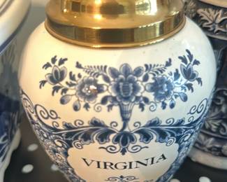 Virginia Blue White Ginger Jar — Decorative blue-and-white ginger jar marked "VIRGINIA" on the front with a domed brass-tone lid. Porcelain-style body features floral and foliate transfer patterns in cobalt blue over a white ground; the lid shows light surface wear consistent with display. Good solid decorative condition with no visible chips or cracks in the photos; ideal as a dining-room accent or mantelpiece. Measures a compact size suitable for tabletop display.