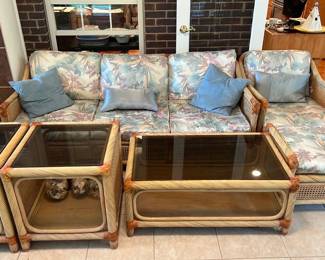 Rattan Patio Set 6pc — Three-piece rattan wicker seating set with matching glass-top coffee and two end tables, upholstered with floral pastel cushions. The woven rattan frame shows a natural finish with wrapped corner joints and cane panel sides; cushions are removable and covered in a pink/blue/tan floral fabric with a couple of solid blue throw pillows included. Good overall used condition with normal light wear to cushions and minor scuffs to rattan visible in photos; glass tops appear intact. Ideal for sun room or casual living spaces and sold as the complete pictured grouping.