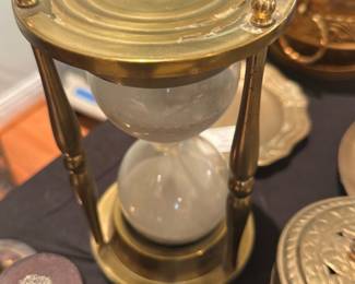 Brass Hourglass Sand Timer — Brass hourglass sand timer with turned column supports and clear glass bulbs. The solid brass frame shows a warm patina and light surface wear consistent with age; glass bulbs appear intact with fine white sand inside. Decorative finials on the top and a stepped base give it a classic look suitable for desk or mantel display. Measures compactly and is ready to use as a decorative timer or accent piece.