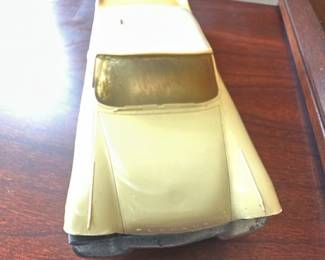Yellow Toy Car Model — Metal toy car model in pale yellow with a white roof and gold-tinted windows; photographed in the office room. Solid die-cast style construction with metal chassis, simple axles and hubcap-style wheels; appears to be a 1950s–1960s style American sedan replica. Overall compact tabletop size (measurements estimated) and lightweight for easy display or shelf decor. Shows paint wear, scuffs and surface scratches, with one front bumper slightly bent and light tarnish on metal parts; mechanically the wheels roll but one axle shows slight play.