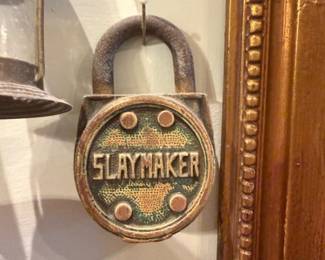 Slaymaker Decorative Padlock — Cast metal padlock with raised "SLAYMAKER" lettering on the face; appears to be an old or reproduction branded lock used decoratively. Heavy iron/bronze-tone finish with verdigris/green patina in recessed areas and four raised rivet details. Shows surface rust and wear consistent with age or outdoor use but no visible broken parts; shackle intact. Useful as a display piece, hardware collectible, or reproduction architectural accent; measures small/handheld size as shown.