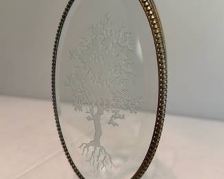 Etched Glass Tree on Metal Stand — Decorative oval etched glass panel featuring a detailed tree and root motif set in a ribbed metal frame with an integrated curved metal stand. The piece appears to be brass-toned metal with light patina and surface wear consistent with age; glass etching is intact with no visible chips or cracks. Stands freely for tabletop display as photographed and makes an attractive accent for a bedroom, shelf, or small tabletop vignette. Solidly made but vintage-condition metal shows expected tarnish that could polish if desired.