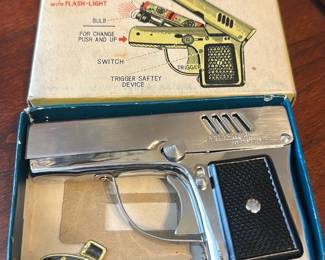 Aurora's Pistol Lighter with Flash-Light — Aurora's pistol-shaped lighter with built-in flash-light is a novelty collectible presented in its original illustrated box. The chrome-tone metal lighter features a black textured grip, trigger-style actuator, and a small bulb flashlight compartment as shown on the box art; the original box shows operating diagrams. Overall compact size suitable for display on a shelf or in a collector's case. In good condition cosmetically with light surface wear to metal and minor edge wear to the box; functionality untested so sold as collectible/parts or display.