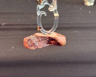Glass Bird Figurine on Stone Base — A delicate clear glass bird figurine mounted on a small natural stone base. The piece features a U-shaped glass form with a stylized bird head and beak accented by small gold-colored details; the stone base has an earthy, reddish tone. Ideal as a small shelf accent or part of a glass-figurine collection; shows light surface dust and typical display wear but no obvious chips or cracks visible in photos.