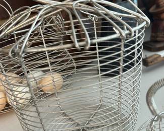 Wire Egg Basket with Handle — Lightweight wire egg basket with a spring-top closure and looped handle; the open-wire construction keeps contents visible and ventilated, making it practical for kitchen or farm use. Construction appears to be nickel- or chrome-plated steel with some light surface wear but no obvious breaks in the wire; closure springs and handle are intact. A simple utilitarian piece that works for storing eggs, produce, or as rustic decor; measures compactly and stacks with similar baskets. Sold as shown.