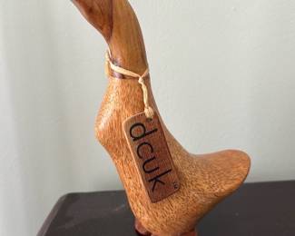 dcuk Wooden Duck Carving — dcuk wooden duck carving with original dcuk hang tag and branded round dcuk mark on the base. Hand-carved from warm-toned wood with a smooth, lightly polished surface and simple painted eyes; the head and body show straightforward folk-style carving. Good decorative condition with minor surface wear consistent with age and display; no cracks visible in photos. Compact accent piece that works well on a bookshelf, mantel, or bedside table.