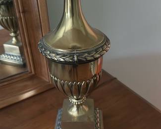 Brass Table Lamp with White Shade — Ornate brass-finish table lamp with a classic urn form and decorative laurel banding, shown with a white fabric bell shade. The metal body features aged highlights and carved-style details on the base and finial, giving a traditional, neoclassical look that suits a bedroom or entry chest. Overall height with shade is approximately 28 inches and the weighted metal base offers stability for tabletop use. Good used condition with light surface wear and patina consistent with normal use; shade shows light creasing but no major stains or tears.