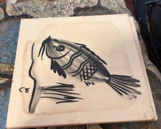 Ceramic Tile — Hand-painted ceramic tile featuring a monochrome fish sketch in ink-like brush strokes on a cream background. The tile has a felt or cardboard backing applied and shows light surface wear and minor staining consistent with age and display; no visible cracks or chips on the face. Lightweight decorative tile suitable for tabletop display, trivet use, or framing; measures roughly 6 inches square (see dimensions).